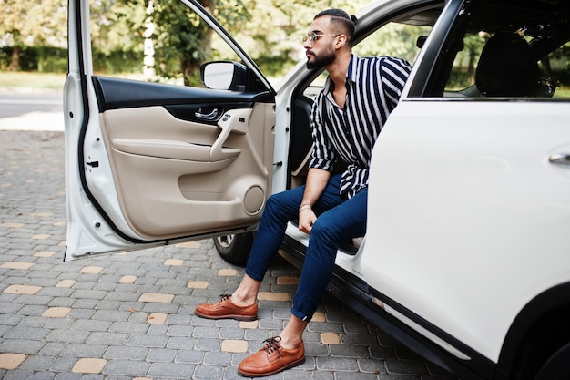 L'uomo arabo di successo indossa una camicia a righe e occhiali da sole pongono al volante della sua auto bianca suv eleganti uomini arabi in trasporto