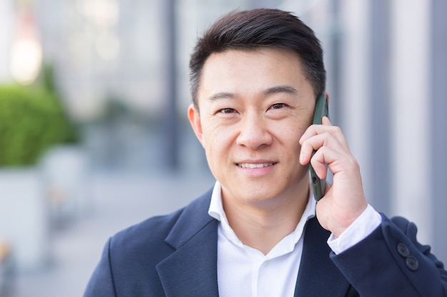 Successful asian businessman businessman talking on cell phone while walking near big modern office, cheerful smiling worker and looking at camera