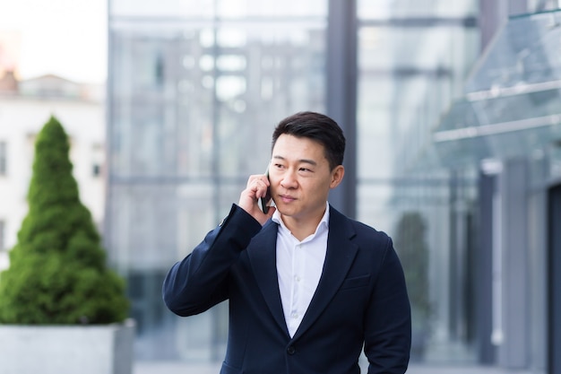 Successful Asian businessman businessman talking on a mobile phone while walking near the office of a large modern, serious and pensive employee