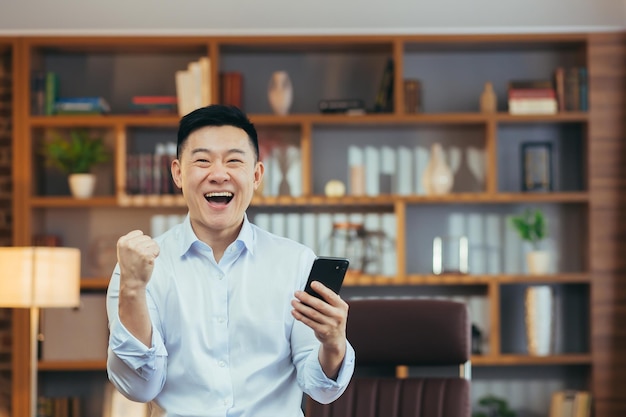 Successful Asian businessman celebrates victory and success man looks at camera and rejoices holds phone win works in classic home office