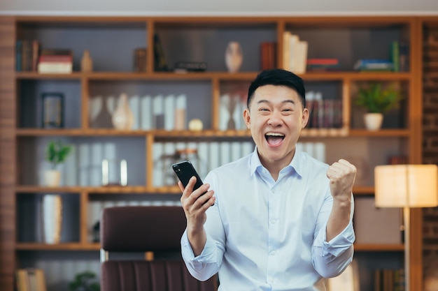 Successful asian businessman celebrates victory and success man\
looks at camera and rejoices holds phone win works in classic home\
office