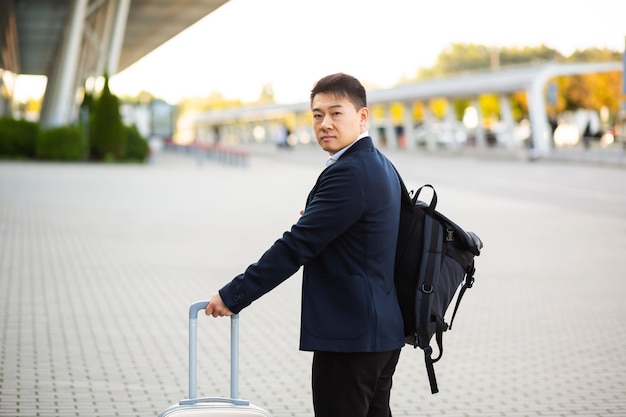 Успешный азиатский бизнесмен возле аэропорта и автовокзала идет с чемоданами серьезного туриста, смотрящего в камеру