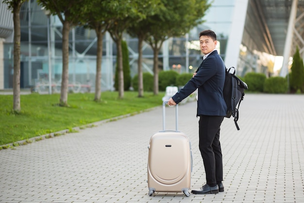 Un uomo d'affari asiatico di successo vicino all'aeroporto e alla stazione degli autobus va con le valigie un turista serio che guarda la telecamera