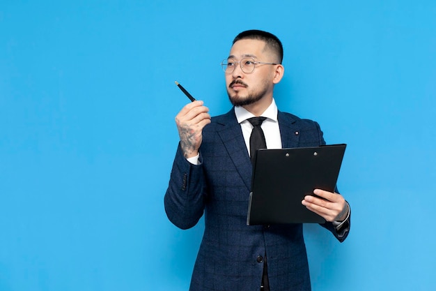 Successful asian businessman in suit and glasses on blue isolated background holds clipboard