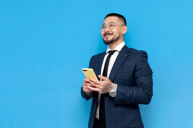 Successful asian businessman in suit and glasses uses smartphone on blue isolated background