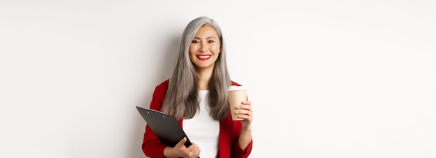 Successful asian businesswoman drinking coffee and smiling standing with clipboard wearing red blaze