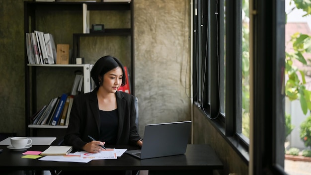 Успешная азиатская деловая женщина сосредотачивается на своей работе на своем ноутбуке в офисе