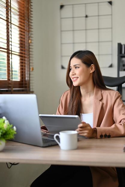 Successful Asian businesswoman at her office desk looking through the window daydreaming