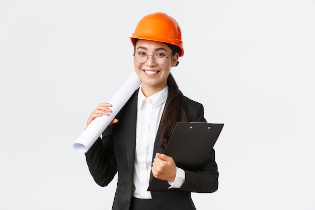 Successful asian female architect in business suit and helmet holding blueprints and clipboard with notes, inspector looking at construction works, smiling at camera, white background