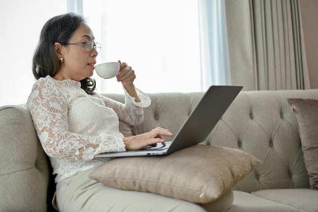 성공한 아시아 중년 여성 사업가가 거실에서 노트북을 사용하면서 커피를 홀짝이고 있다