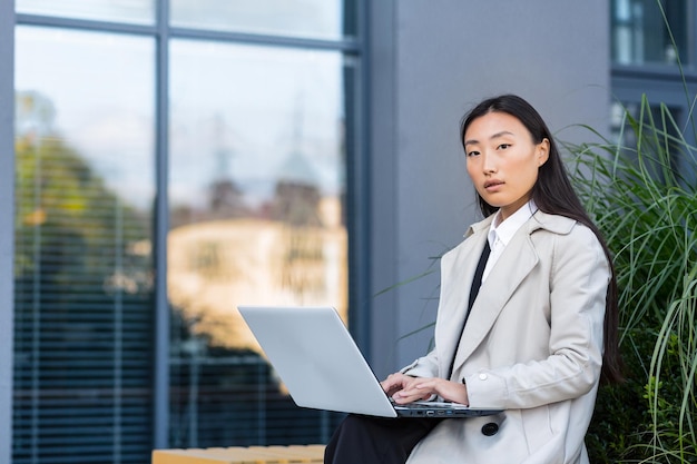 사무실 센터 근처에 앉아서 카메라를 바라보며 야외에서 노트북 작업을 하는 성공적인 아시아 여성 사업가