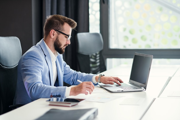 Successful bearded businessman in glasses and in blue suit works with laptop in modern office.