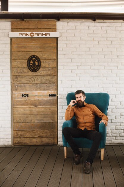 Successful bearded man in a turquoise chair on the terrace near\
a stack of firewood