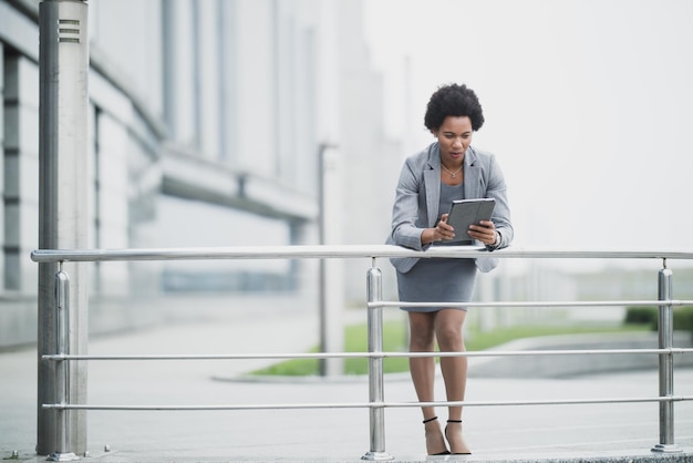 A successful black business woman using app on a digital tablet in front a corporate building.