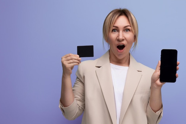 Successful blond woman in jacket with card and phone mockup on studio background