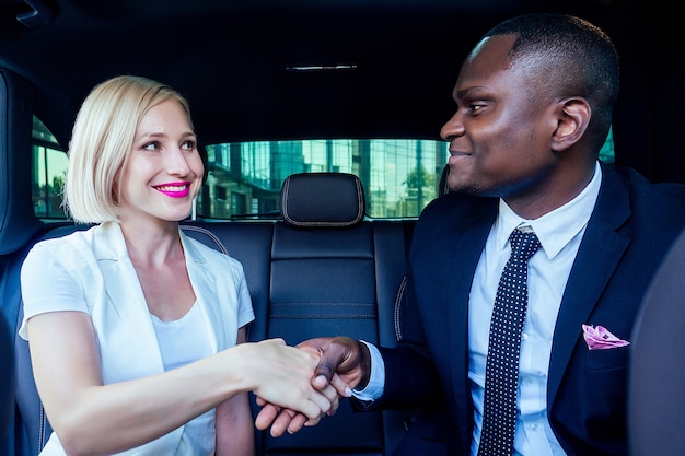 Successful blonde business woman entrepreneur employer with makeup in white dress with a handsome Afro-American boss man industrialist in black stylish suit jacket handshake working in car good deal
