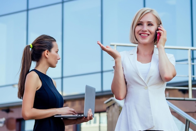 Successful blonde business woman talking on the phone and brunette secretary female person holding file folders and looking at securities laptop in the hands skyscraper background