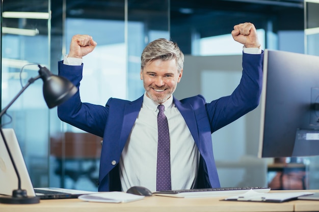 Successful boss works in a modern office at the computer a senior man gray and happy with a beard looks at the camera and smiles holds his hands up celebrates victory