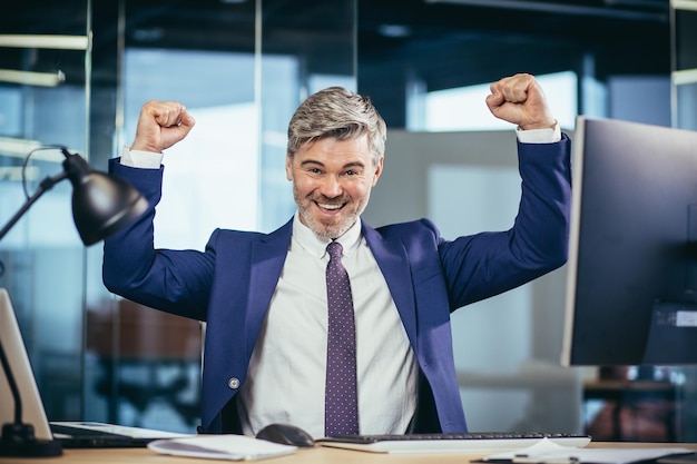 Successful boss works in a modern office at the computer a\
senior man gray and happy with a beard looks at the camera and\
smiles holds his hands up celebrates victory
