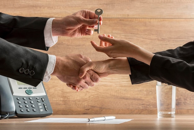 Successful business deal - real estate agent and new female homeowner exchanging house key while shaking hands over a contract of house sale.