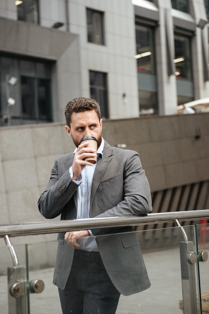 successful business man in gray suit looking aside, while standing and drinking takeaway coffee in front of modern business center