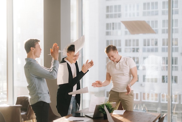 Successful business team are having fun throwing paper documents celebrate corporate success in the sunlit conference room on the background of glass skyscrapers