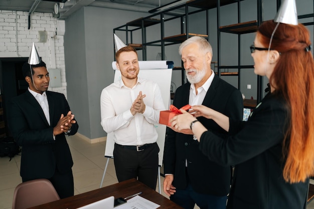 Successful business team of multiethnic people giving present to happy mature adult male boss Multinational businessteam wearing festive holiday hats applauding to mature adult male leader at office