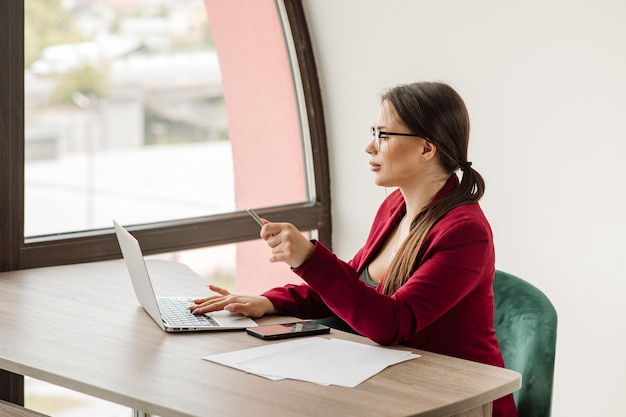 Успешная деловая женщина в красной куртке работает с ноутбуком в офисе