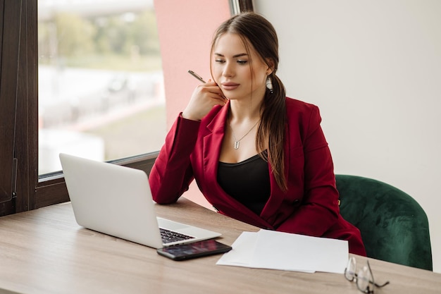 Successful business woman dressed in red jacket is working with a laptop in the office