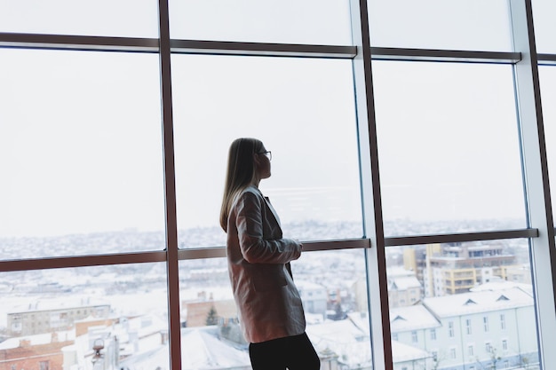 A successful business woman in trousers and a jacket stands and looks out the huge panoramic window of the business center skyscraper