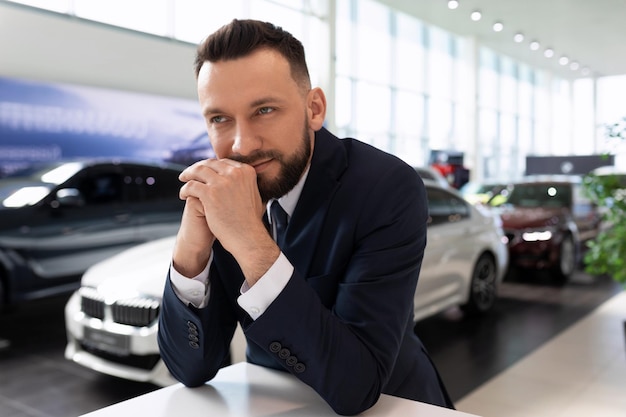 L'uomo d'affari di successo in una concessionaria di automobili accanto a nuove auto guarda pensieroso in lontananza