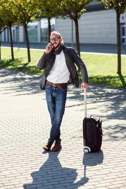 Successful businessman in casual suit pulls a suitcase, hurries to the airport and talks on smartphone. Young caucasian man going on a business trip.