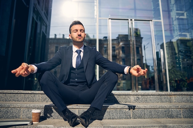 Successful businessman in formal clothes relaxing alone outdoors on stairs while drinking coffee and dreaming