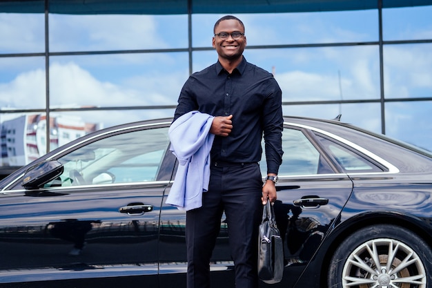 Successful businessman handsome African American man in a stylish suit in a blue jacket black shirt and glasses standing in front of a cool new black car on the street