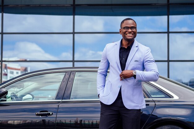 Successful businessman handsome African American man in a stylish suit in a blue jacket standing in front of a cool new black car on the street