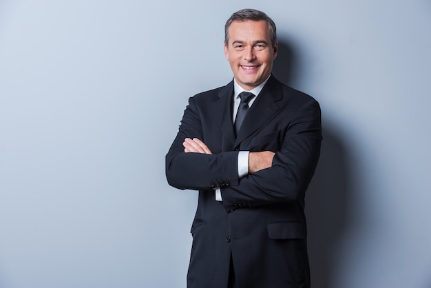 Successful businessman. Portrait of confident mature man in formalwear looking at camera and smiling while keeping arms crossed and standing against grey background