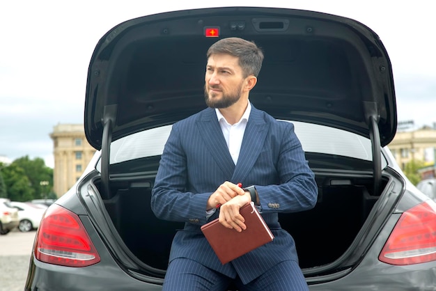 L'uomo d'affari di successo siede con un diario sul cofano della sua prestigiosa auto