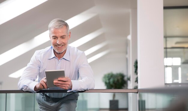 Successful businessman standing at railing in his office, using digital tablet