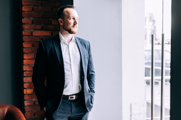 Successful Businessman in a suit stands in an office and looks out through the glass doors.