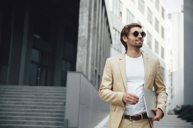 Successful businessman walking near modern dark building and carrying wireless laptop