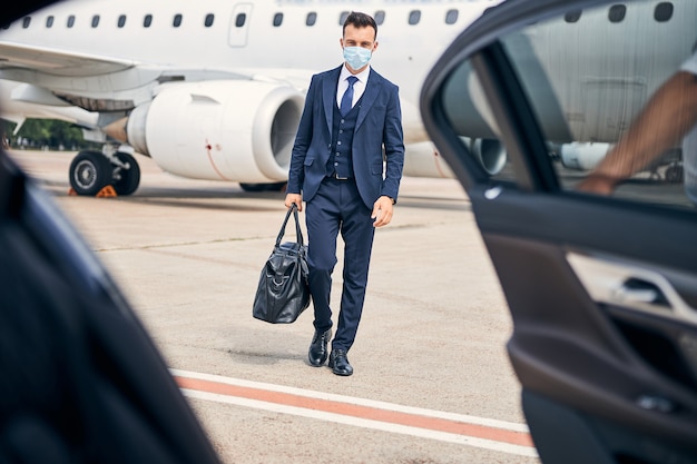 Successful businessman wearing a facial mask walking towards a car