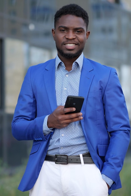 Successful businessman with black smartphone stands and smiles at camera against office building