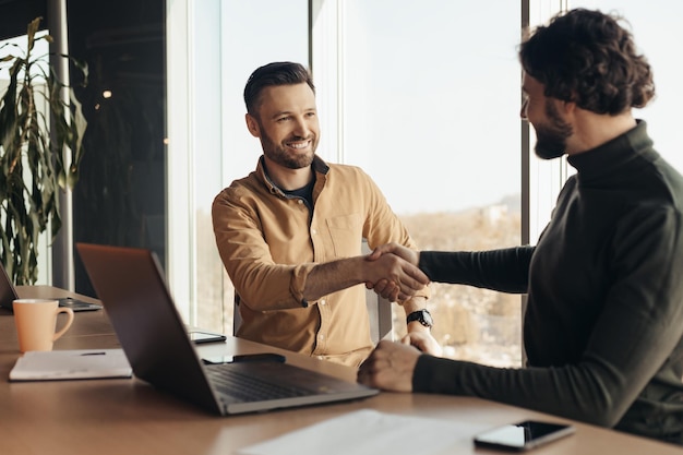Успешные бизнесмены пожимают друг другу руки в знак согласия, сидя за столом с ноутбуком в офисе компании бесплатно