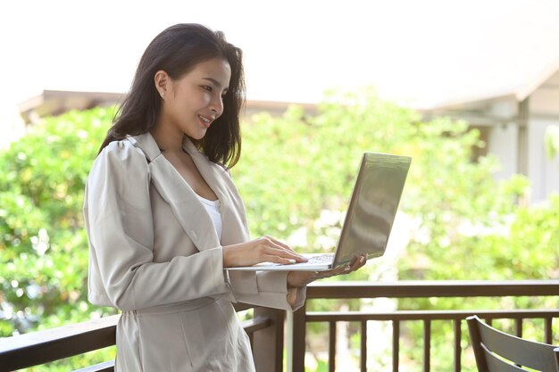 Successful businesswoman standing on a terrace outside office and using laptop