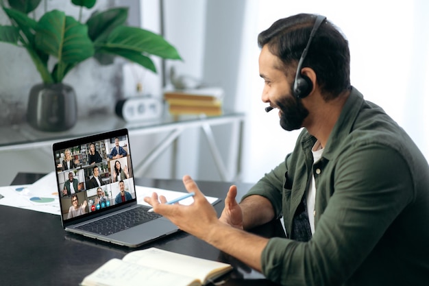 Uomo di successo di successo di razza mista impegnato seduto alla scrivania in un ufficio moderno utilizzando il laptop che parla con un team aziendale multirazziale tramite videoconferenza riunione online virtuale negoziazioni a distanza