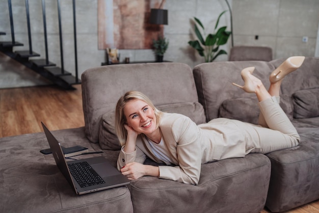 Successful caucasian young businesswoman in beige business suit laying on couch using laptop smiles
