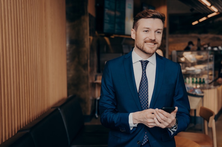 Photo successful cheerful businessman in stylish classic outfit using smartphone while standing in cafe or restaurant, being happy after receiving message with positive news, looking aside and smiling