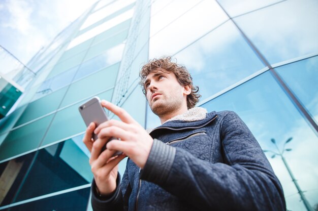 Successful confident handsome attractive young man in black jacket using mobile phone standing near glass building