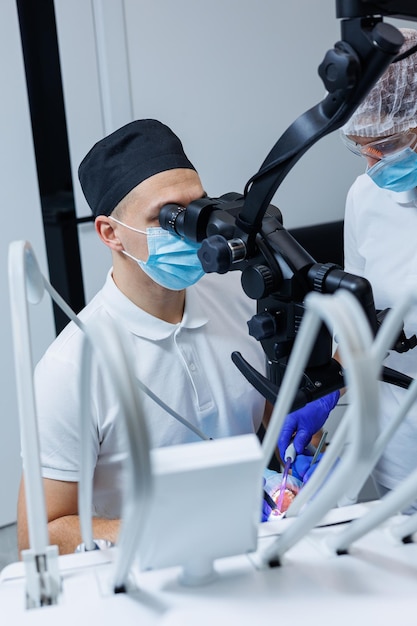 A successful dentist looks at the patients teeth with a dental microscope and holds dental instruments near his mouth The assistant helps the doctor They wear white uniforms with masks and gloves