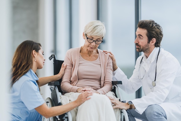 Successful doctor and young nurse talking with their senior female patient in a wheelchair at the hospital hallway.